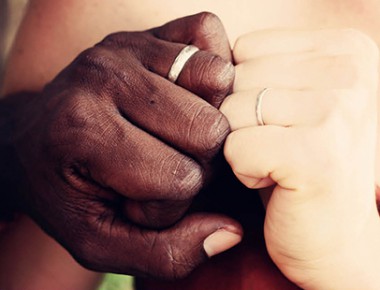 Two hands held close together, wearing wedding bands. The two hands have contrasting skin tones, one dark, one light.