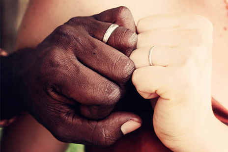 Two hands held close together, wearing wedding bands. The two hands have contrasting skin tones, one dark, one light.